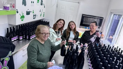 ארבע נשות משפחת היקב מחייכות סביב שולחן העבודה ביום ביקבוק השיראז, מאות בקבוקים על הדלפק