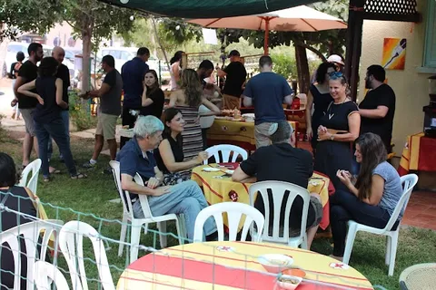 אורחים יושבים סביב שולחנות עם מפות צבעוניות על הדשא בצל העצים בחצר יקב סודות