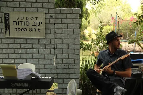 נגן גיטרה בכובע יושב ליד שלט יקב סודות באירוע ההשקה בחצר היקב